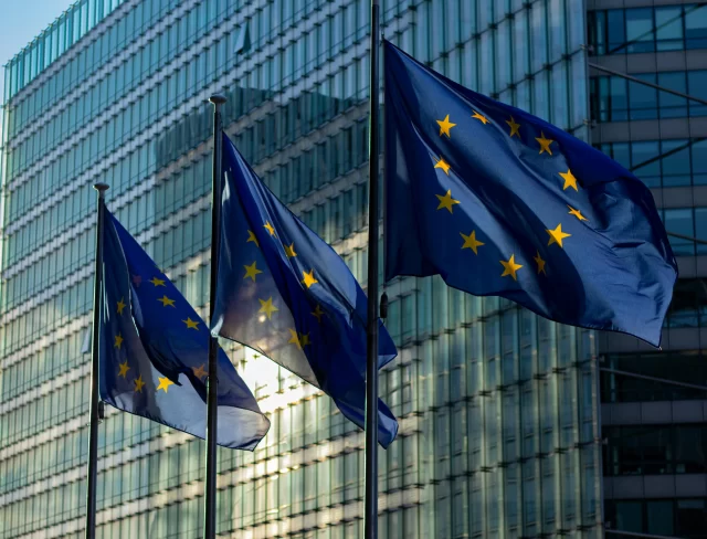 Three European Union flags waving in front of a glass building, representing the EU context of the ePrivacy Regulation.