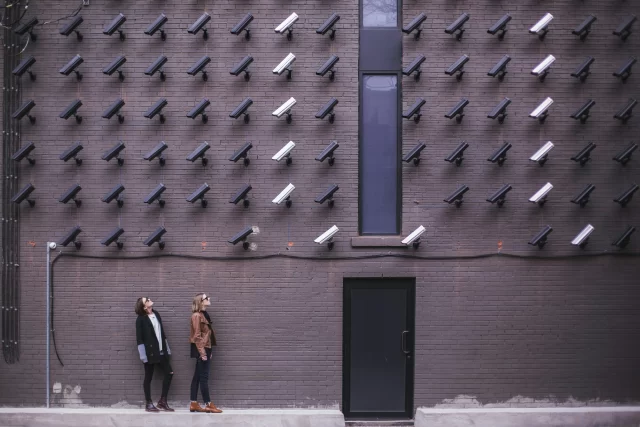 Two women standing before a wall of cameras, illustrating the intense surveillance involved in maintaining PCI data security.