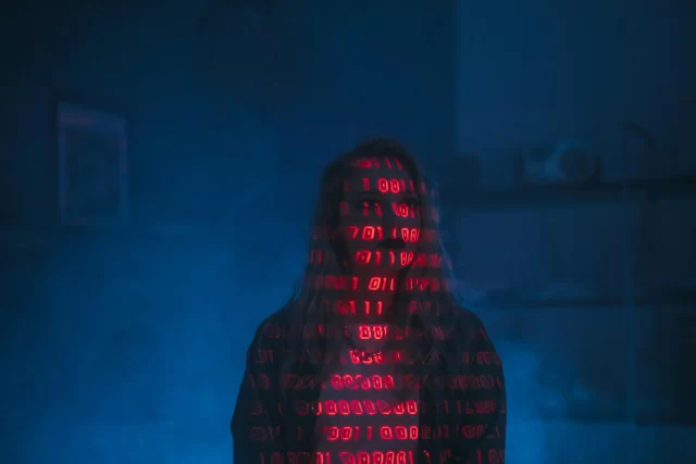 A woman standing in a dark, hazy room with binary code projected across her face and body, symbolizing a Data Breach.
