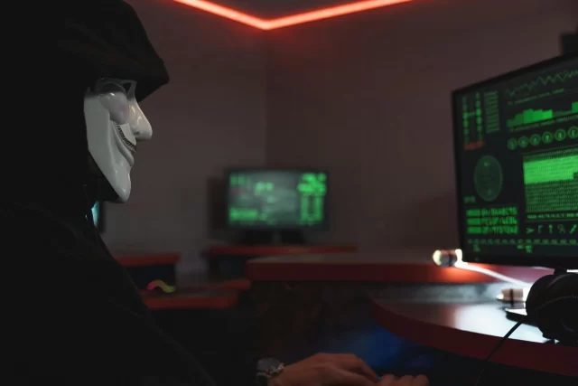 Person in a Guy Fawkes mask at a computer terminal, illustrating the threat of Database Breaches.